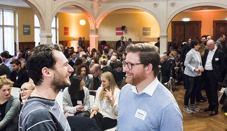 Zwei Herren unterhalten sich im gefüllten Saal. Im Hintergrund zwei sich austauschende Damen und weitere Gespräche