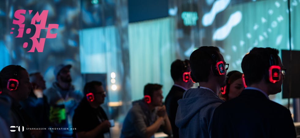 Besucher der Symbioticon tragen leuchtende Over-Ear-Kopfhörer in illuminiertem Raum