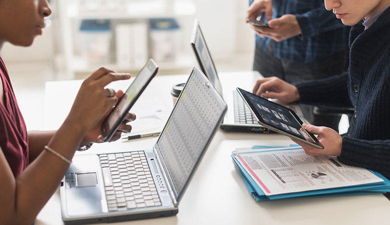 Moderne Büroarbeiter sitzen an einem gemeinsamen Tisch, einer steht, es wird auf Smartphones, Tablets und Laptops geschaut