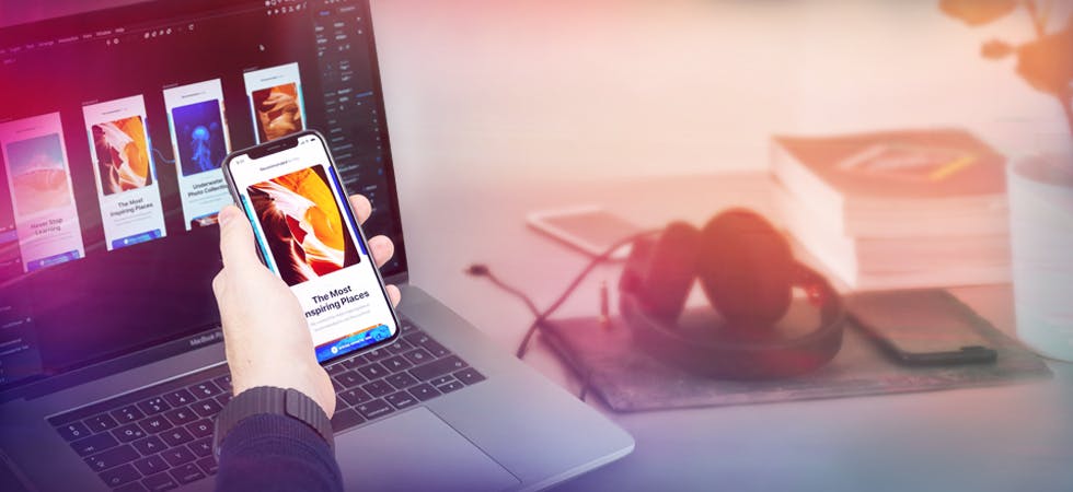 User hält sein Smartphone vor seinen Laptop, im Hintergrund Kopfhörer