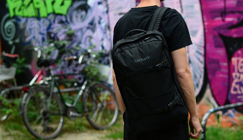 Ein junger Mann steht mit dem Rücken zum Betrachter, der Rucksack im Fokus. Im Hintergrund Fahrräder vor einer mit Graffiti besprühten Wand
