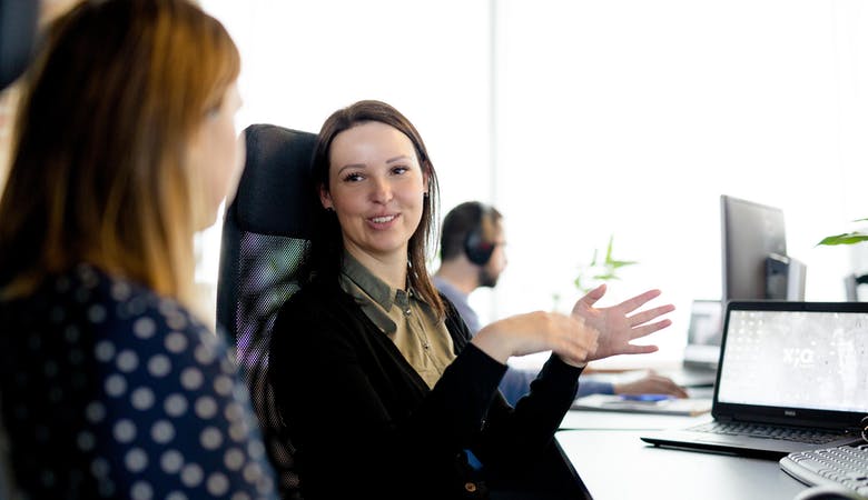 Zwei junge Frauen sitzen am Schreibtisch, Laptop im Hintergrund, unterhalten sich