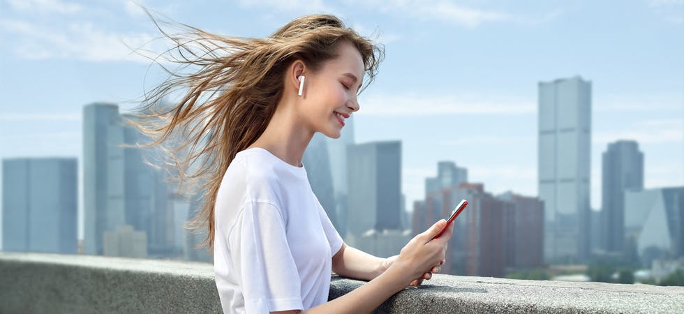Eine junge Frau steht auf einem Hochhausdach an der Reling, mit Bluetooth-Kopfhörern im Ohr und einem Smartphone in der Hand. Sie lächelt