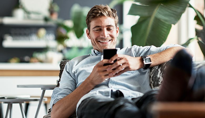 Ein weißer Mann mittleren Alters liegt mit Smartphone in der Hand auf einem Sofa, im Vordergrund ein Blatt einer Zimmerpflanze
