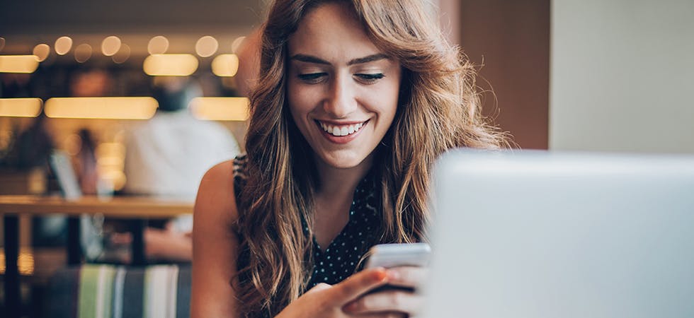 Junge Frau sitzt vor Laptop, schaut aber aufs Smartphone. Im Hintergrund durch Tiefenschärfe verschwommene Lichter und weitere Menschen zu erkennen
