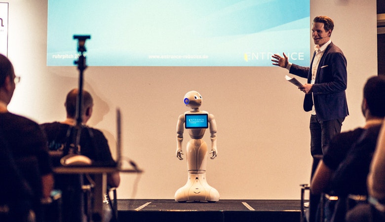 Sebastian Holtze auf der Bühne vor einer Leinwand, neben ihm ein kleiner Roboter mit Bildschirm auf der Brust