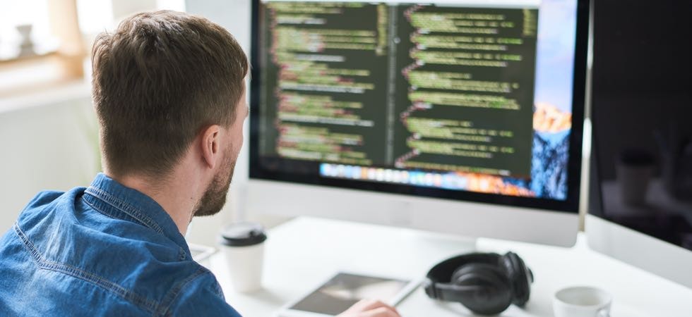 Ein junger Developer mit Kurzhaaarfrisur sitzt im blauen Jeanshemd an seinem Schreibtisch. Darauf ein To-Go-Kaffeebecher, eine Tasse und Over-Ear-Kopfhörer.