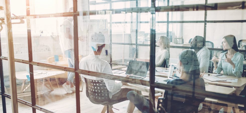 Aufmacherbild des Advertorials über den HubSpot Sales Hub. Zu sehen eine gruppe junger Menschen in einem von Glas umfassten Meetingraum. Ein Speaker steht vor sitzender Gruppe an Tisch, Blicke auf ihn gerichtet.